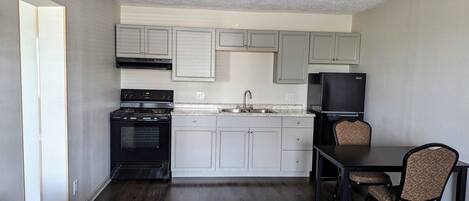 A compact kitchen with a stove, sink, and refrigerator, a dining table with chairs, and a dark wooden floor.