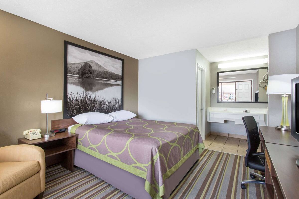 A hotel room with a bed, a desk with a lamp, a chair, a telephone, a bathroom with a mirror, and a window.