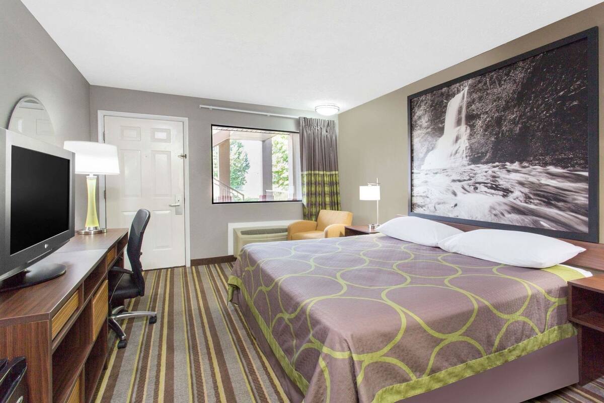 A hotel room with a bed, a desk with a TV, a chair, a window with curtains, and a large framed picture on the wall.