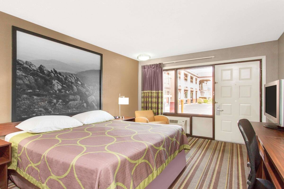 A hotel room with a large bed, a desk with a chair, a television, and a view of the outside through a door.