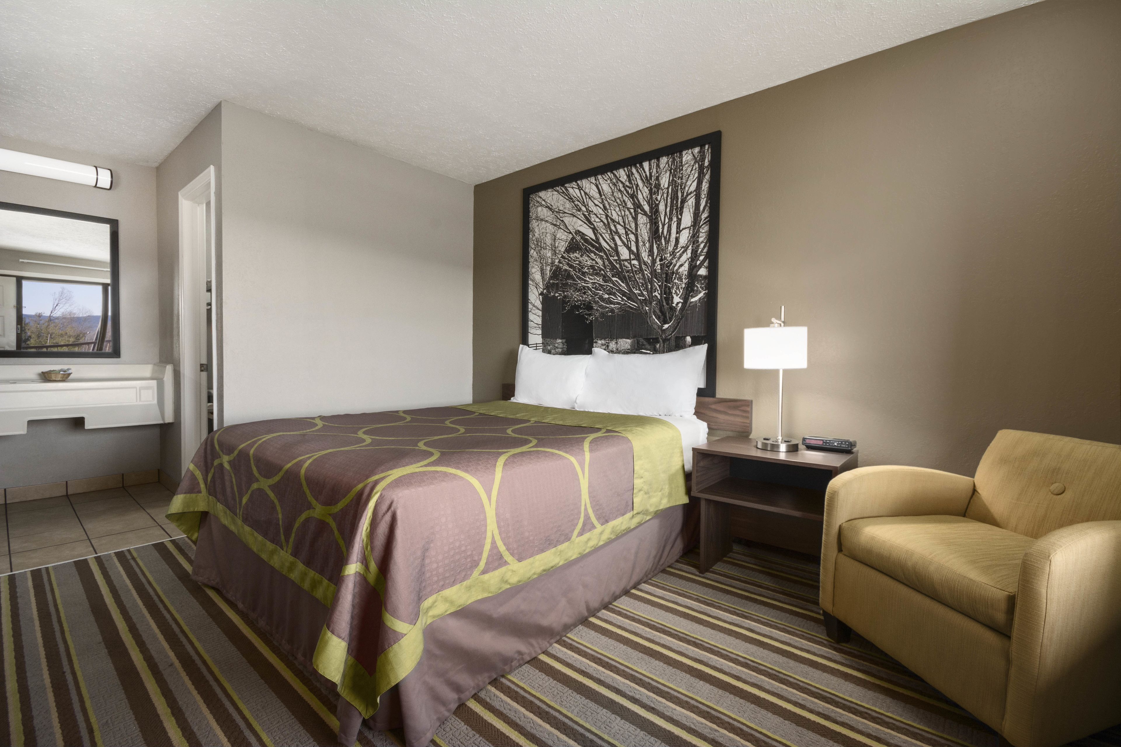 A hotel room with a bed, a chair, a desk, a lamp, and a large framed picture on the wall.