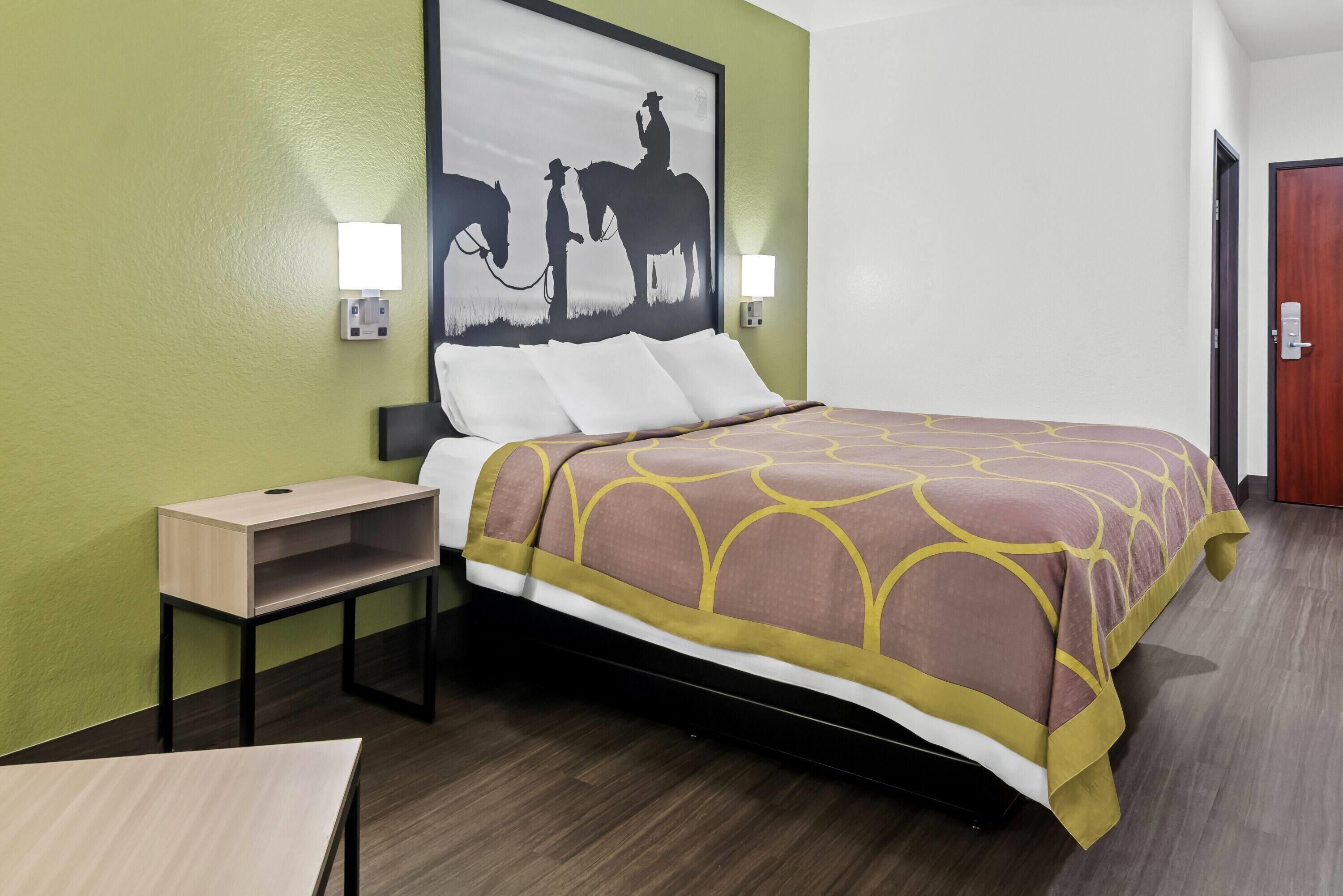 A hotel room with a bed, a nightstand, a table, and a wall-mounted painting of a cowboy and horse.