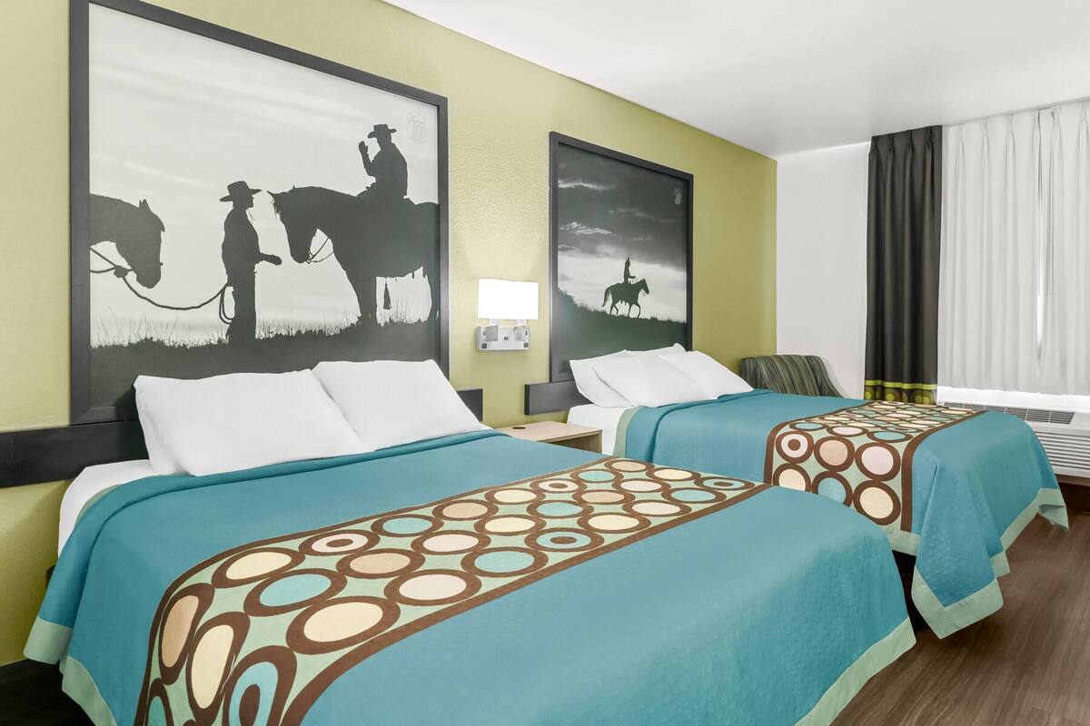 A hotel room with two beds, each with a patterned bedspread, a nightstand with a lamp, and a large framed picture of a cowboy on a horse.