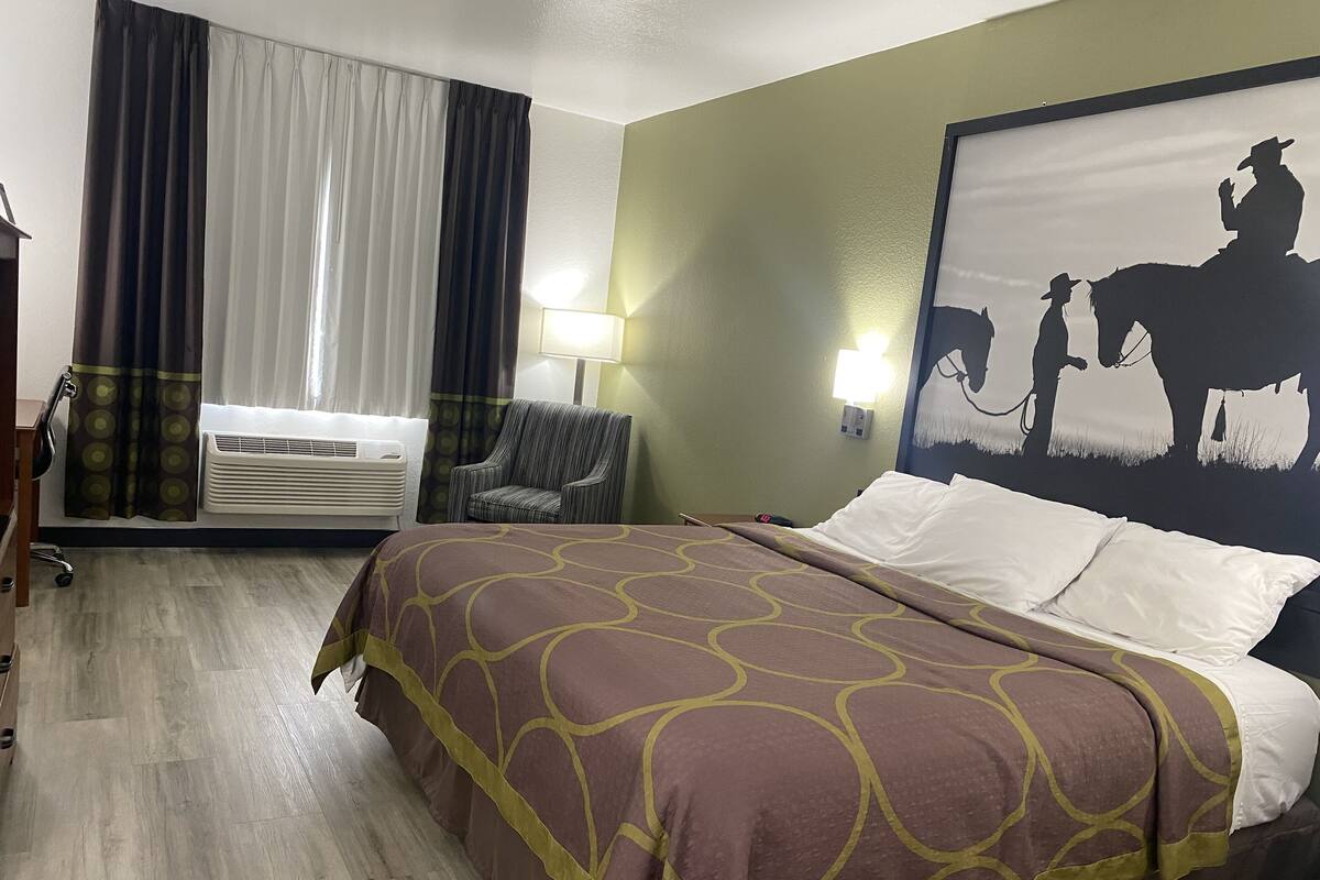 A hotel room with a bed, a chair, a wall-mounted lamp, an air conditioning unit, and a large painting.