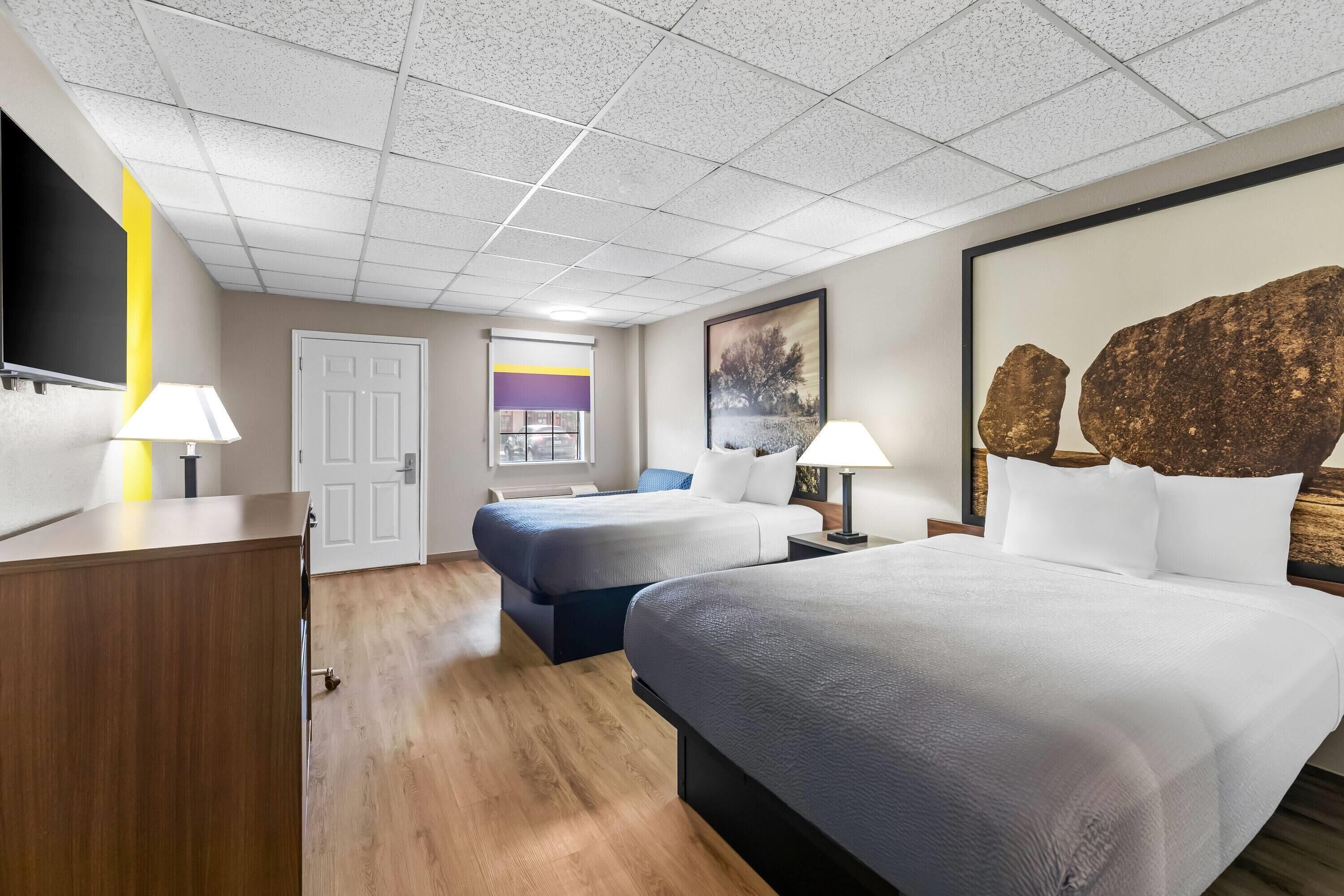 A hotel room with a large bed, two smaller beds, a desk, a TV, and a painting on the wall.