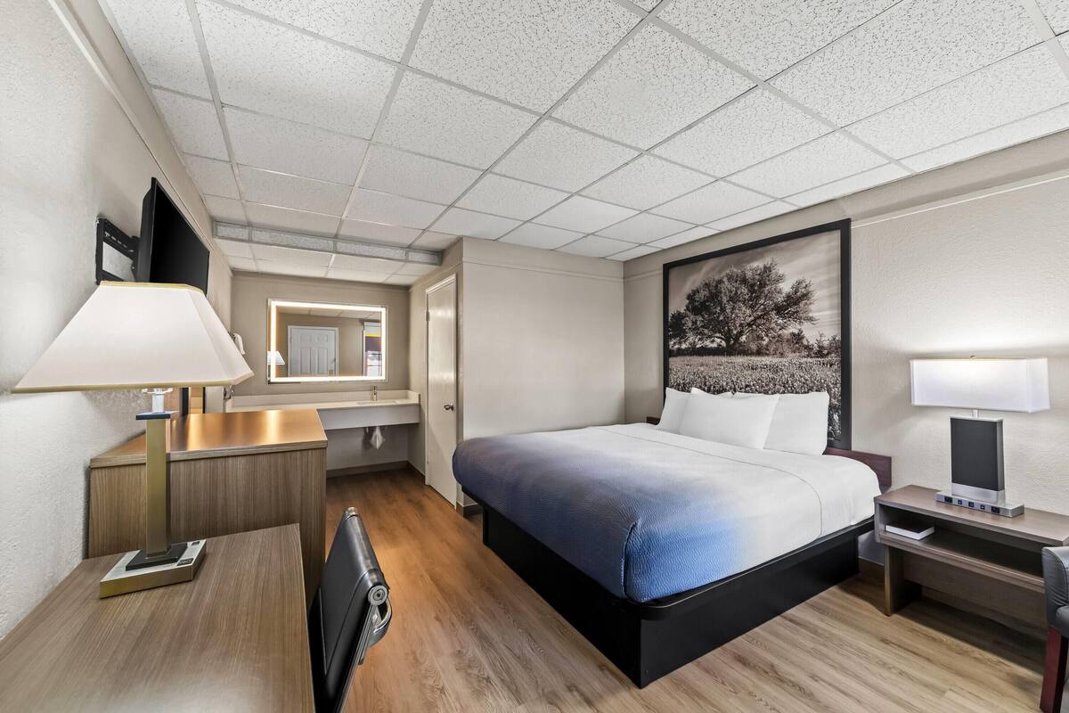 A hotel room with a large bed, bedside tables, a desk, a chair, a desk lamp, a mirror, and a framed picture on the wall.