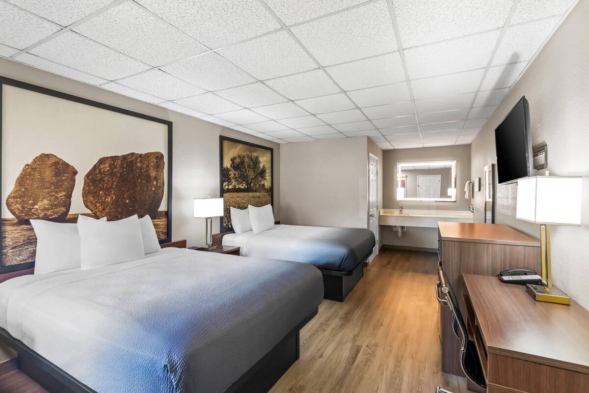 A hotel room with two beds, a desk, a TV, and a bathroom visible in the background.