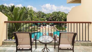 Un balcón con dos sillas de mimbre y una pequeña mesa redonda, con vista a una zona de piscina que incluye sillones de descanso y una estructura con techo de paja.