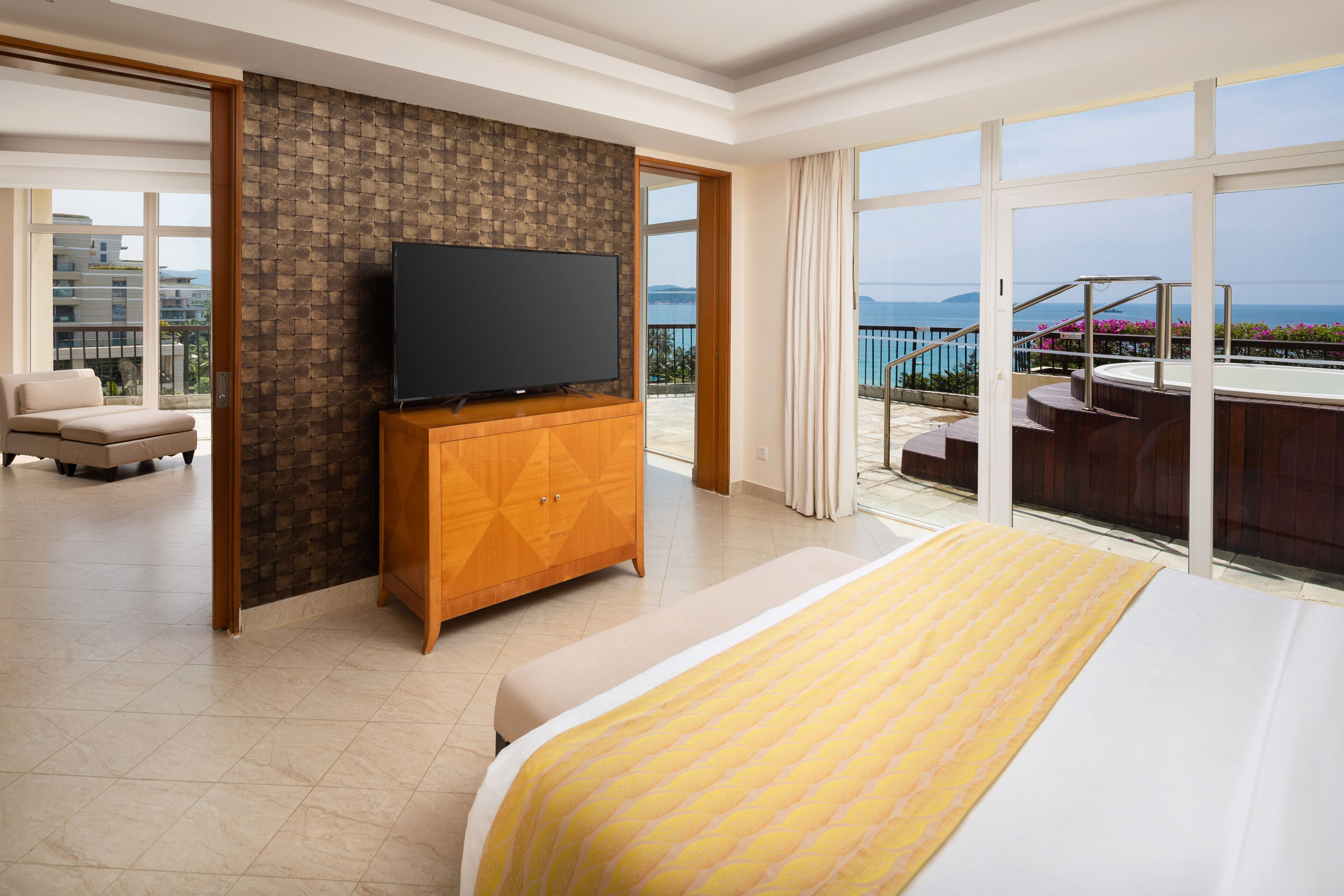 A hotel room with a large bed, a TV on a wooden cabinet, a balcony with a pool, and a view of the sea.