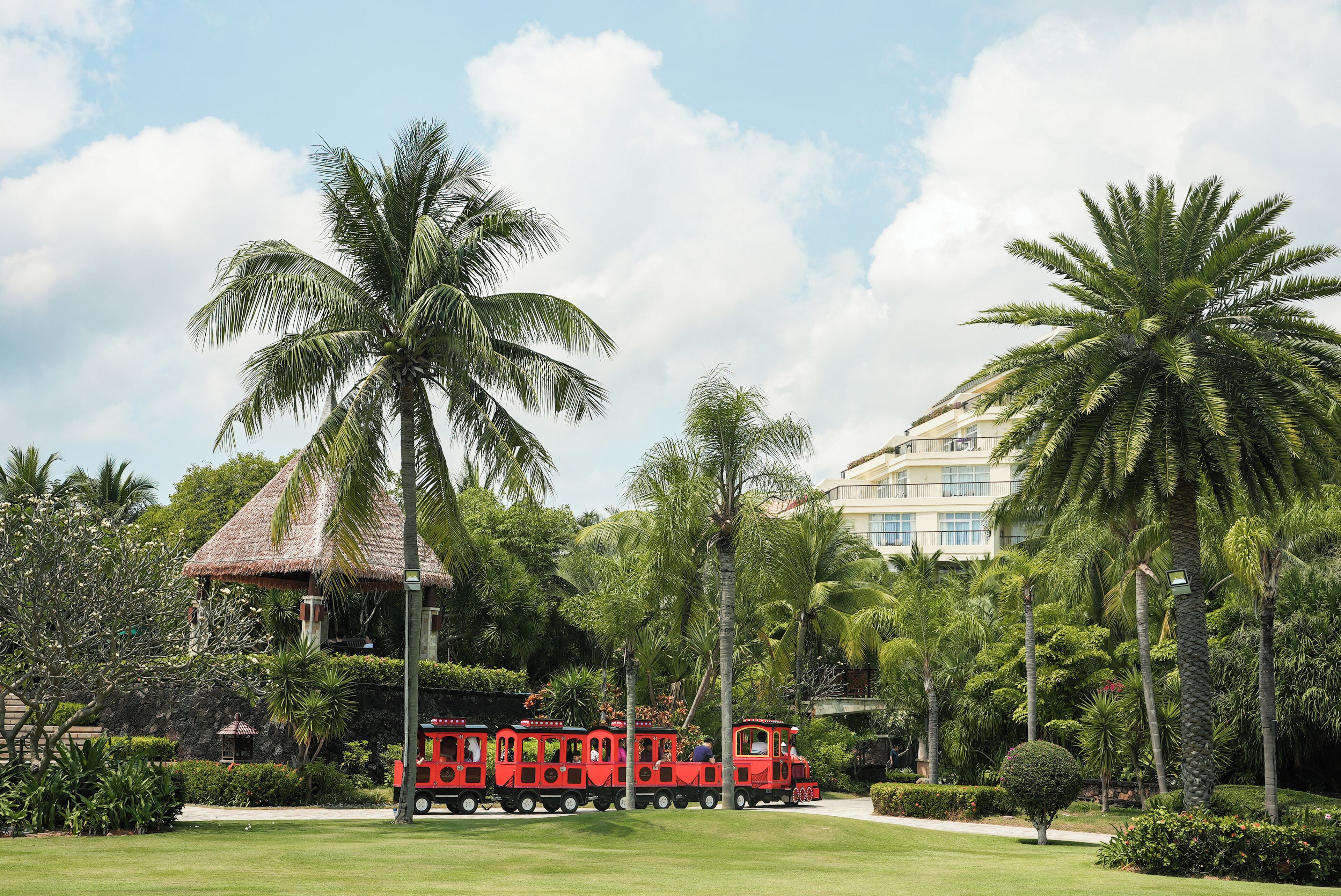 A red train in a tropical garden with palm trees and a building in the background.