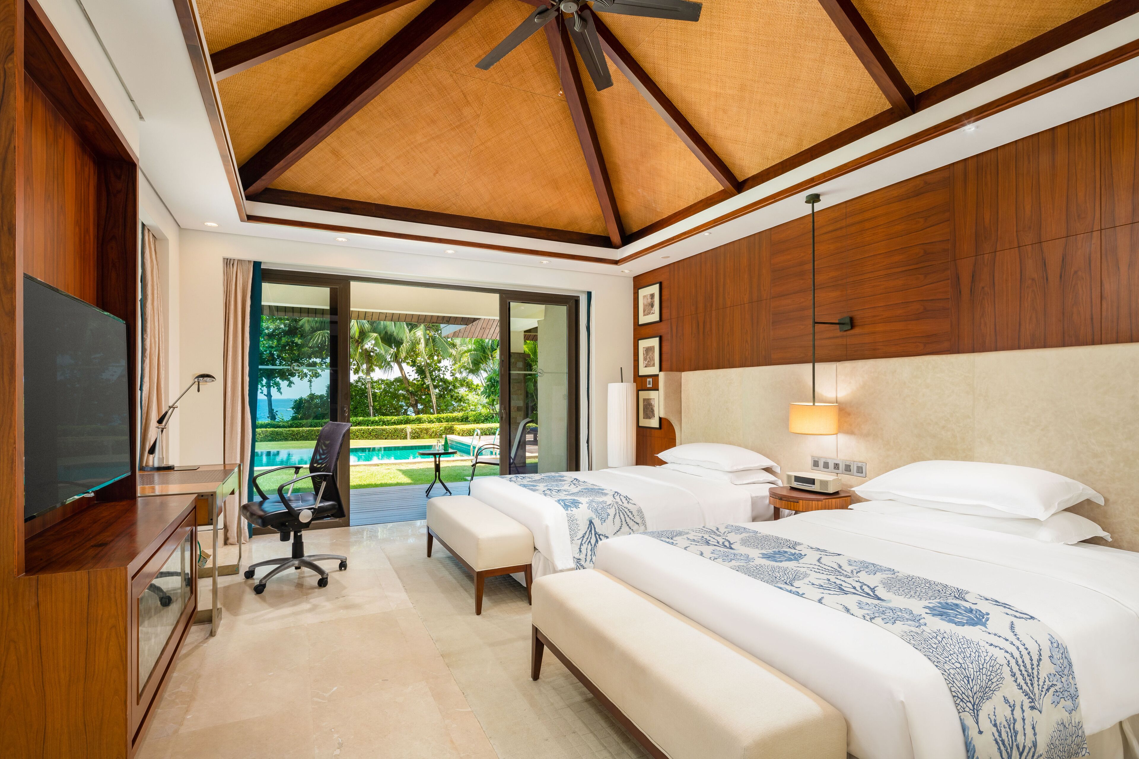 A hotel room with two beds, a desk, a TV, and a balcony with a pool view.