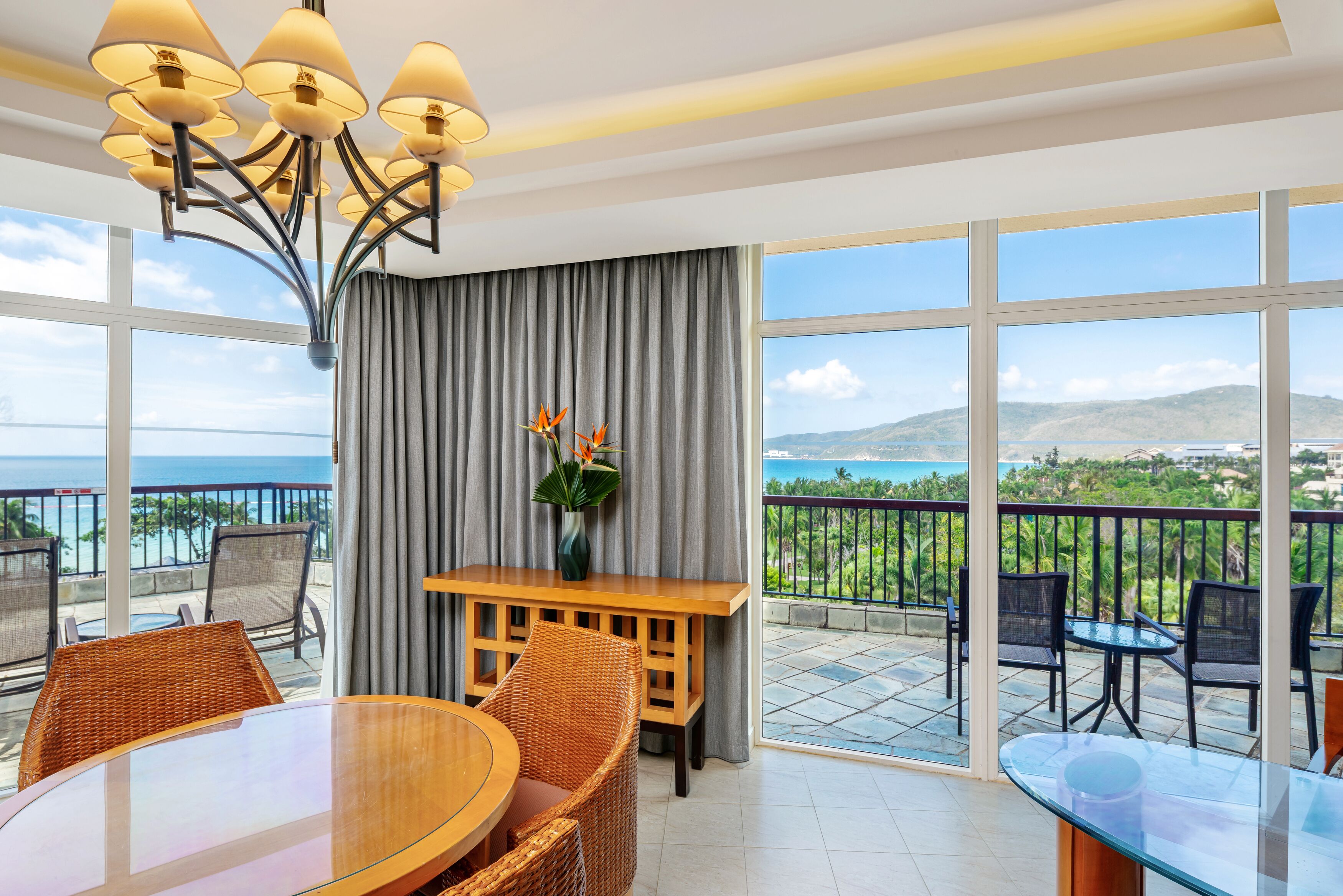 A hotel room with a balcony, a dining table, wicker chairs, a vase with flowers, and a view of the sea and mountains.