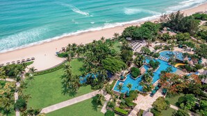 Vista aérea de un complejo turístico con playa, piscina y abundante vegetación.