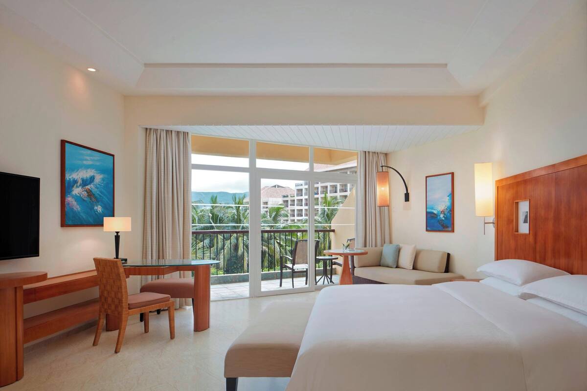 A hotel room with a large bed, a desk, a chair, a sofa, and a balcony with a view of palm trees and buildings.