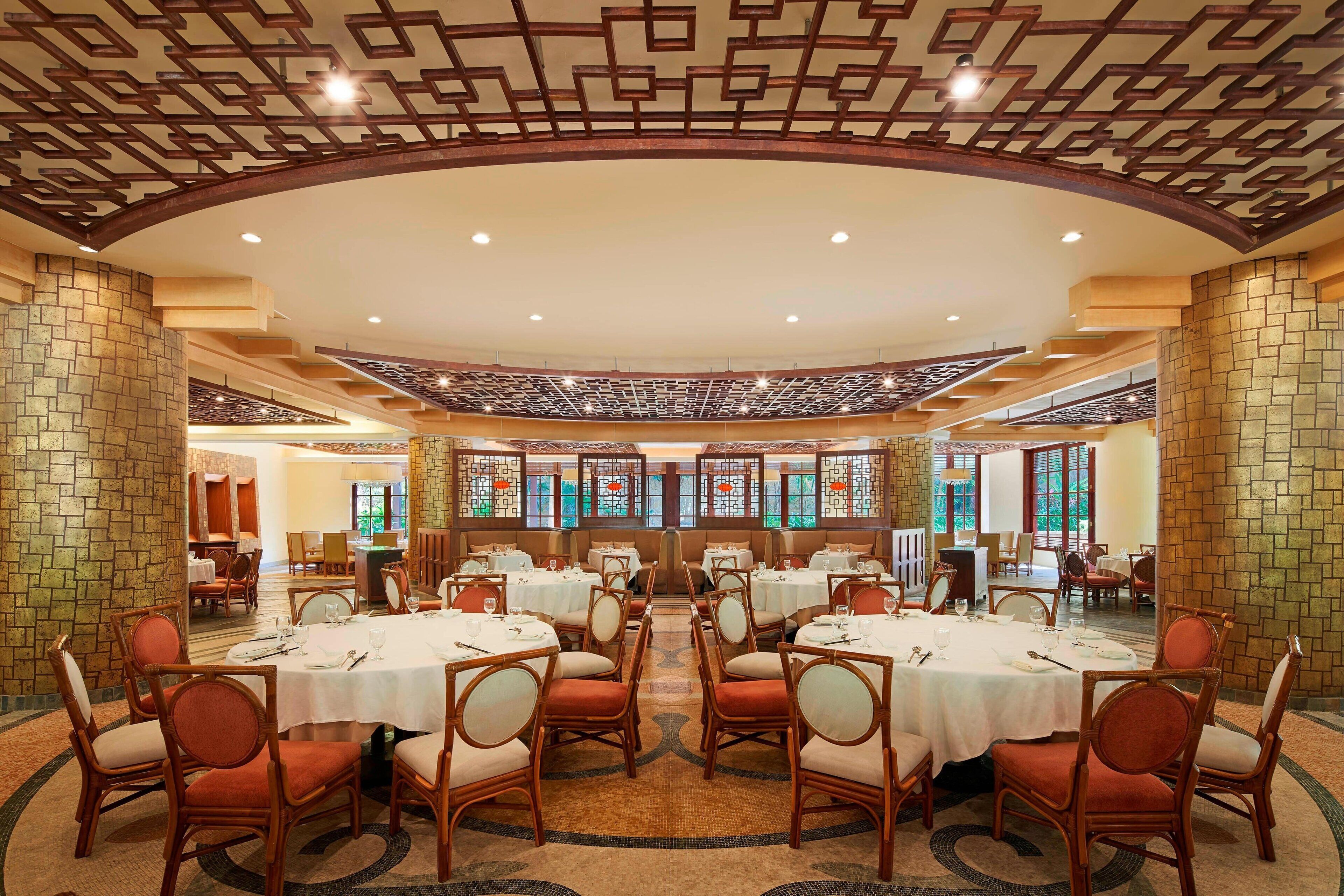 A spacious dining area with round tables and chairs, a decorative ceiling, and large windows with stained glass.