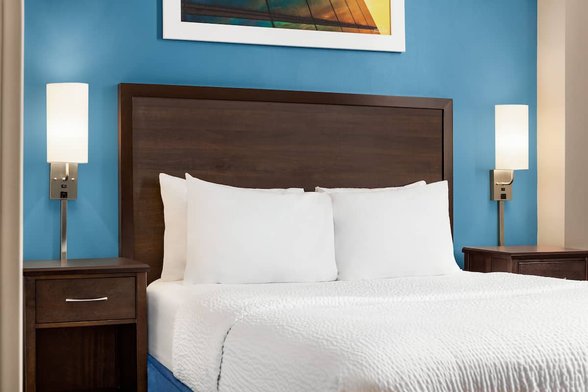 A neatly made bed with white linens and pillows, flanked by two bedside tables with lamps, set against a blue wall with a framed artwork above.