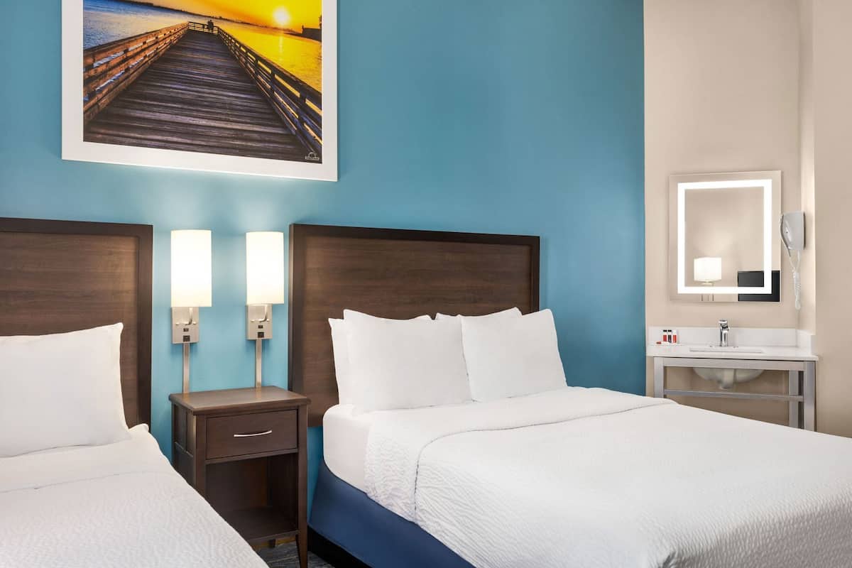 A hotel room with two beds, a nightstand with a lamp, a bathroom with a mirror, and a painting of a pier on the wall.