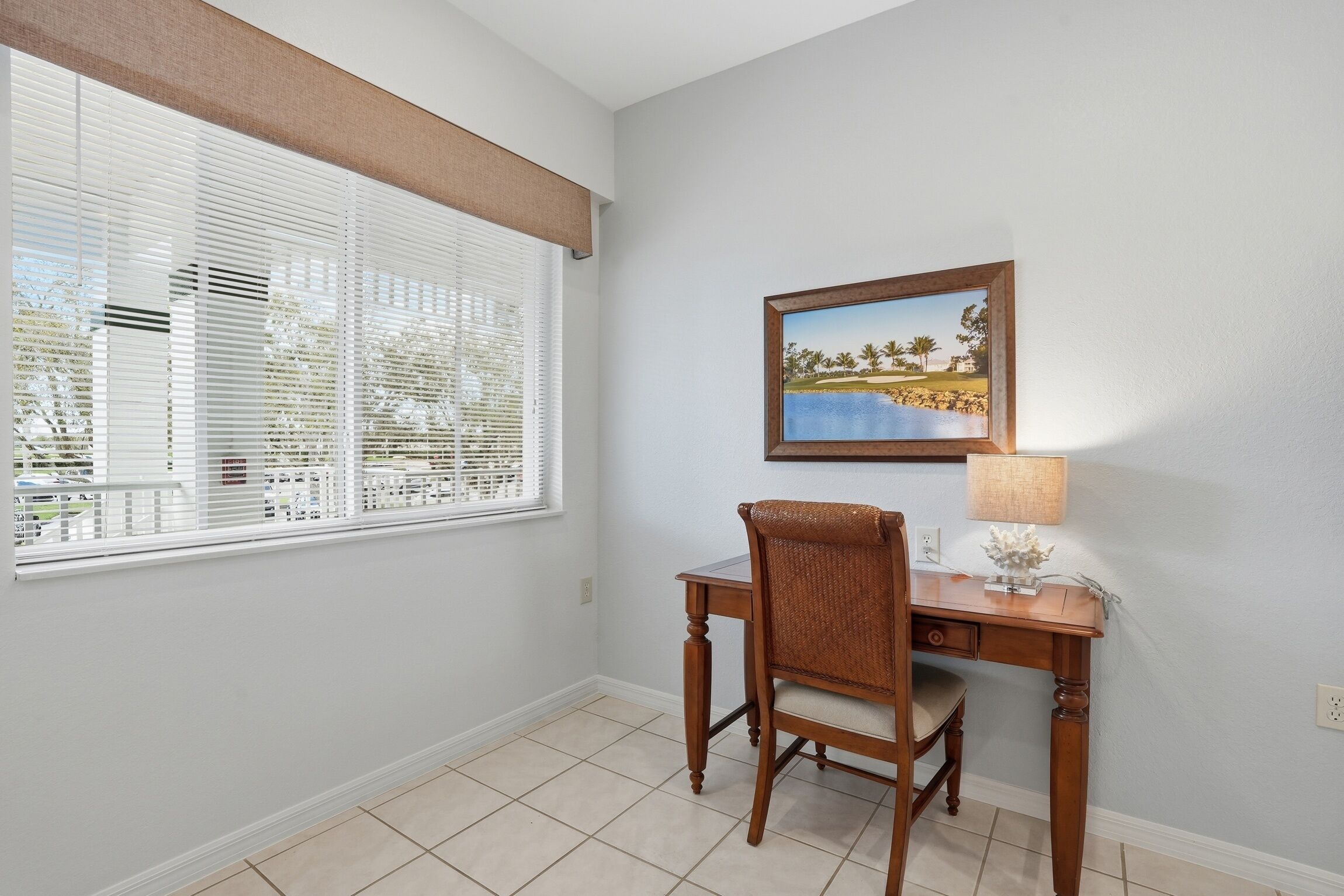 A room with a desk, chair, window with blinds, and a framed picture on the wall.