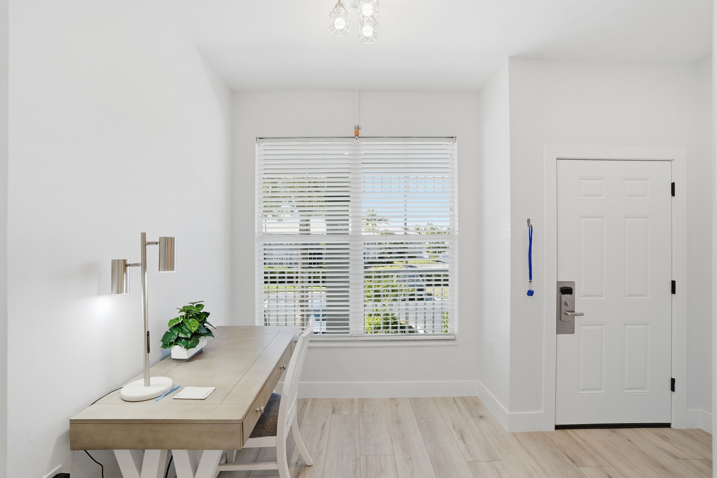 Una habitación con una mesa de madera, una planta en maceta, una puerta blanca y una ventana con persianas.