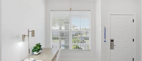 Una habitación con una mesa de madera, una planta en maceta, una puerta blanca y una ventana con persianas.