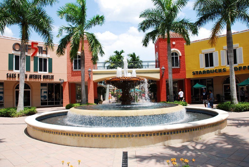 A fountain with water jets in the center of a plaza surrounded by palm trees and storefronts, including a Starbucks.