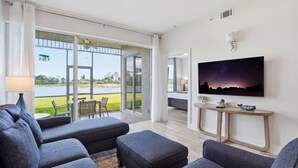Una sala de estar moderna con un televisor de pantalla plana grande, sofás grises, una mesa de centro y vista a un lago a través de amplias puertas de cristal.