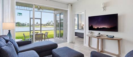Una sala de estar moderna con un televisor de pantalla plana grande, sofás grises, una mesa de centro y vista a un lago a través de amplias puertas de cristal.