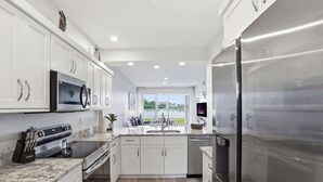 Una cocina moderna con armarios blancos, electrodomésticos de acero inoxidable, encimera de granito y vistas al exterior.
