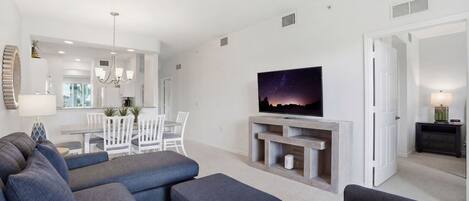 A modern living room with a flat-screen TV, grey sofas, a dining area, and a chandelier.