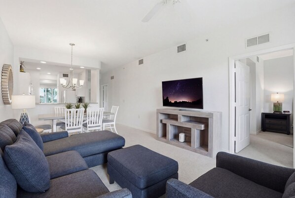 A modern living room with a flat-screen TV, grey sofas, a dining area, and a chandelier.