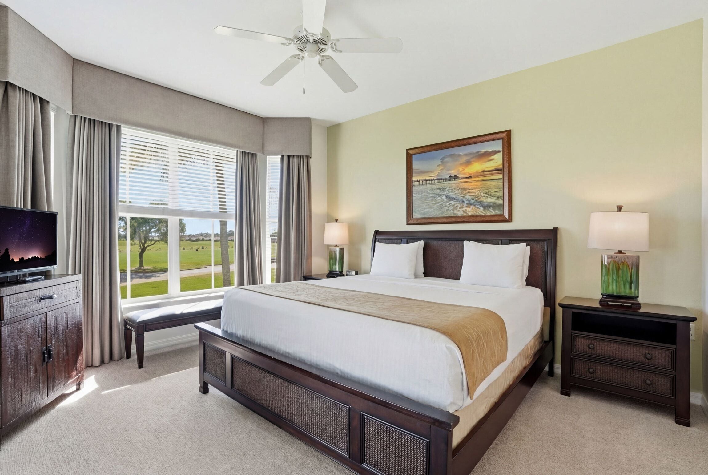 A hotel room with a large bed, a television, a ceiling fan, a painting, and a window with curtains.