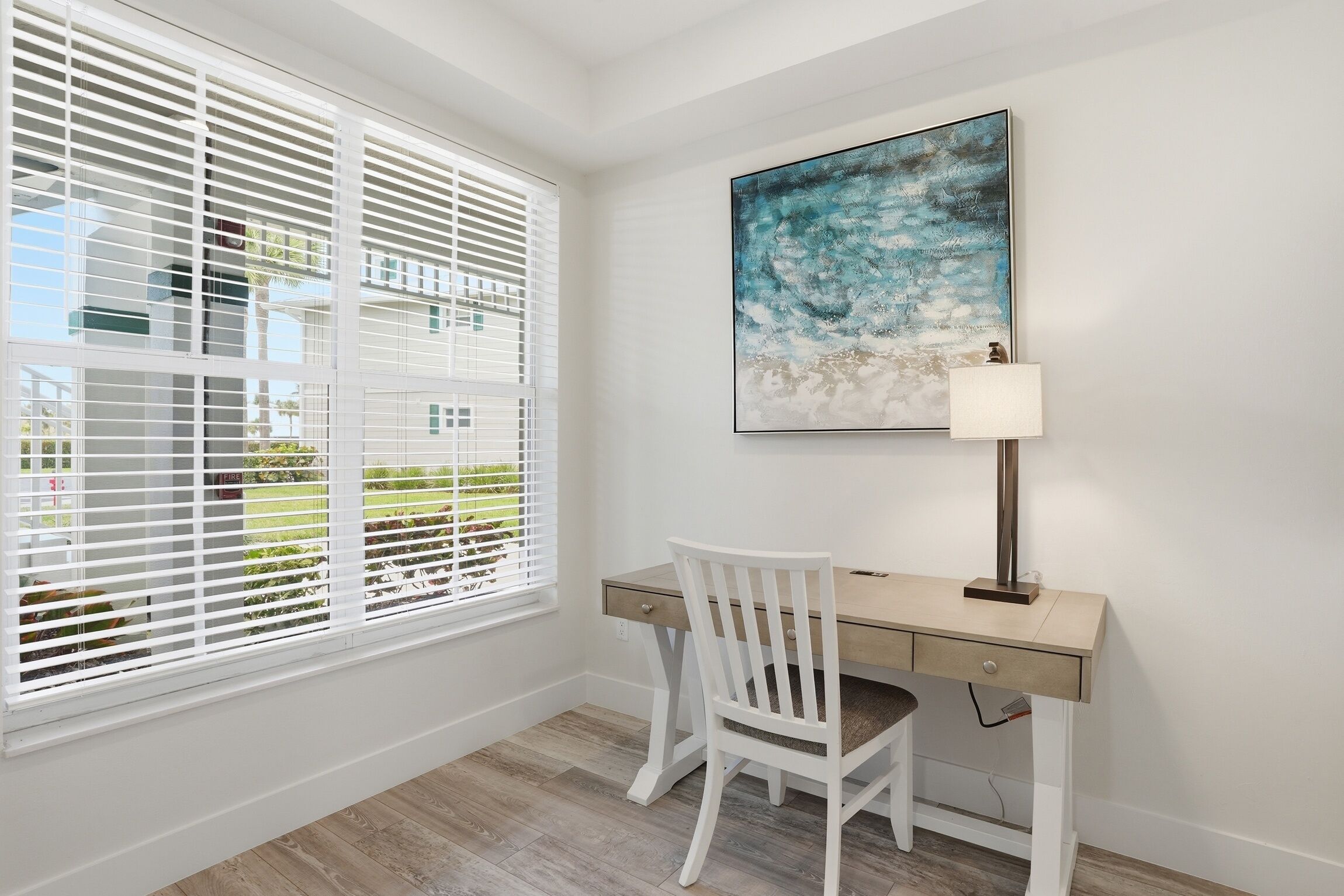 A room with a desk, chair, window with blinds, and a painting on the wall.