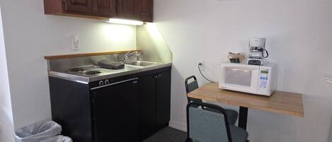 Une petite cuisine équipée avec une cuisinière, un évier et un four à micro-ondes. On y trouve une petite table avec une chaise, et une poubelle est visible.