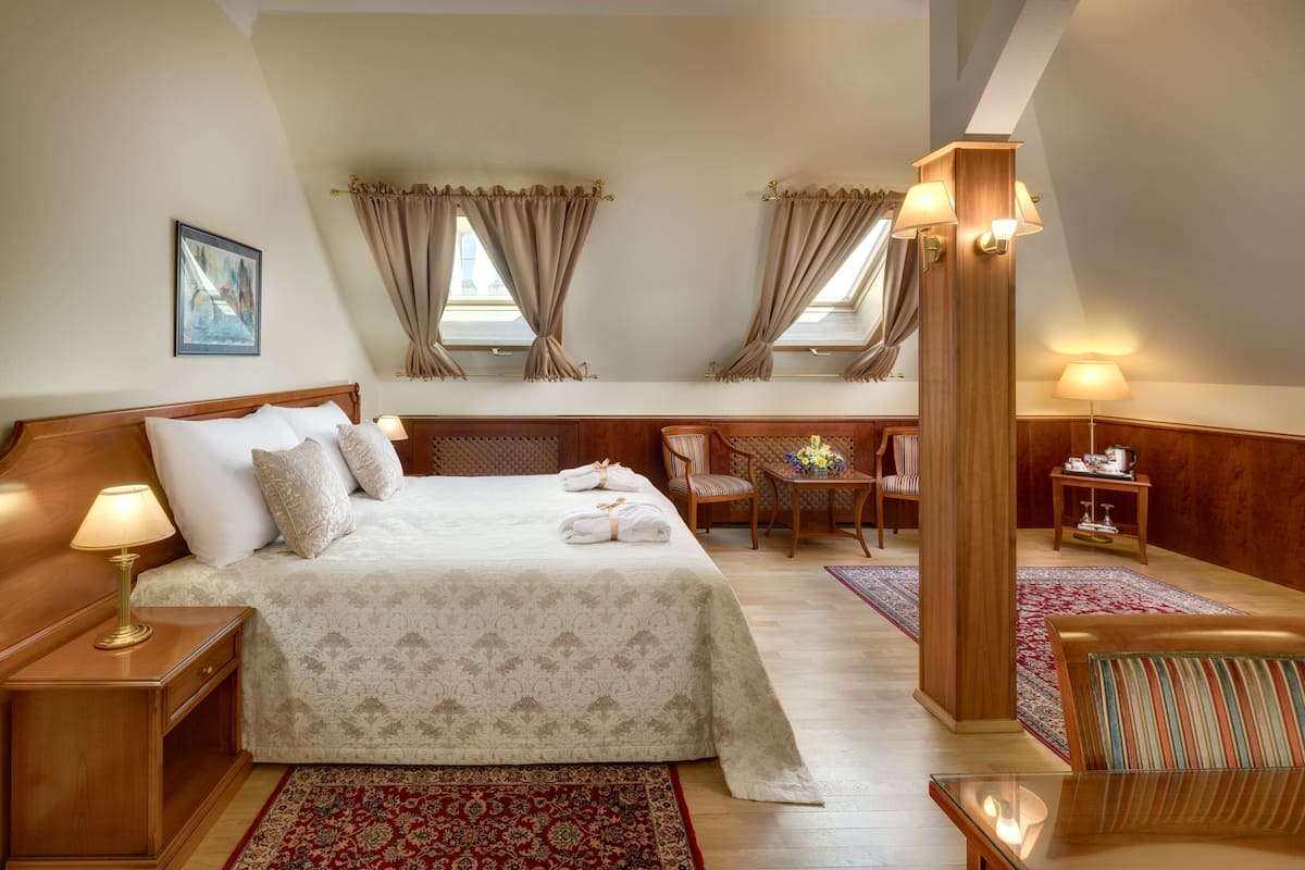 A bedroom with a large bed, a desk, and a chair. There are two windows with curtains, a lamp, and a small table with a vase of flowers.