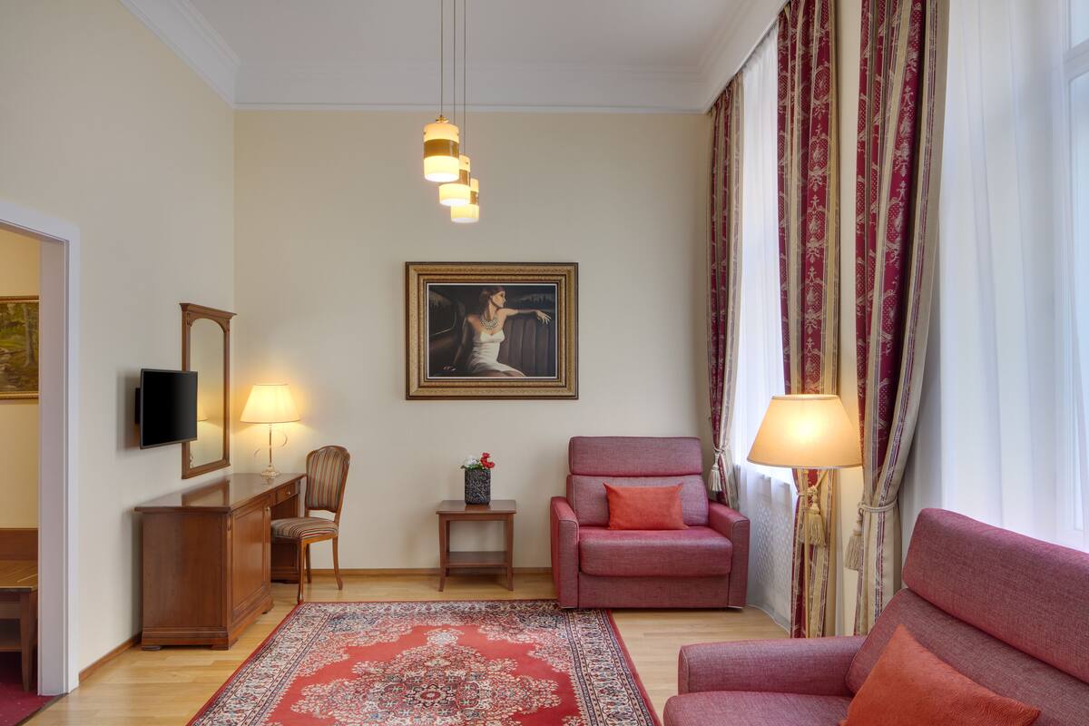 A living room with a red sofa, a smaller armchair, a wooden cabinet, a TV, a framed painting, and a patterned rug.