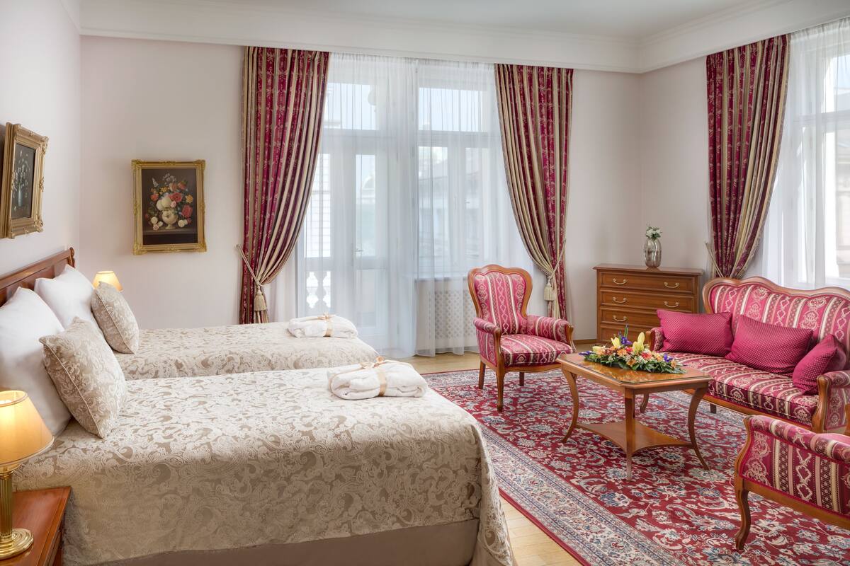 A hotel room with two beds, a sitting area with a sofa and armchairs, a wooden coffee table, and a large window with curtains.