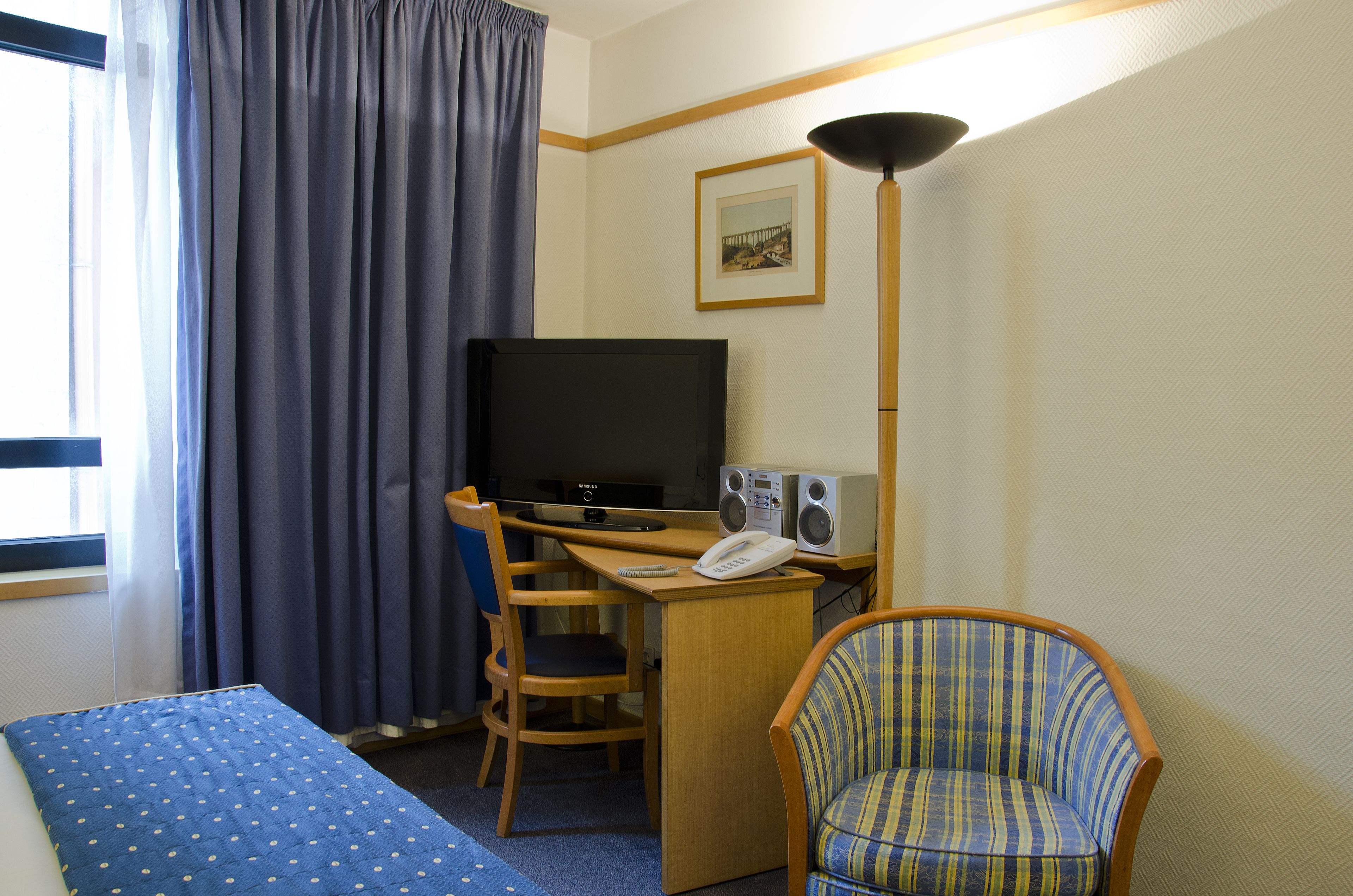 A hotel room with a bed, a desk with a computer and speakers, a chair, and a TV.