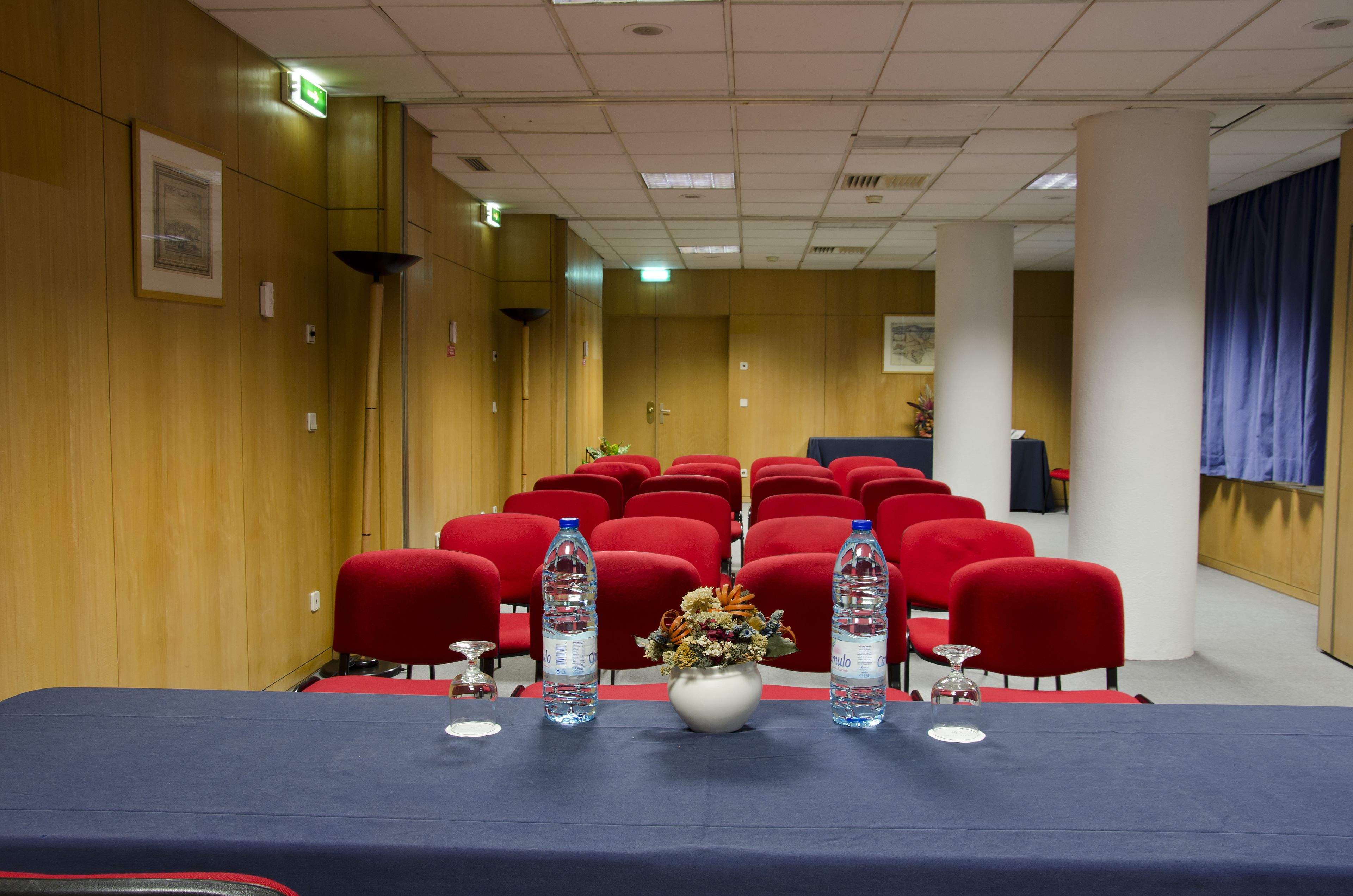 Une salle de conférence avec une longue table, des chaises rouges, des bouteilles d’eau et un vase rempli de fleurs.