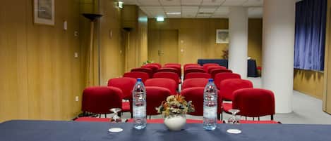 Une salle de conférence avec une longue table, des chaises rouges, des bouteilles d’eau et un vase rempli de fleurs.