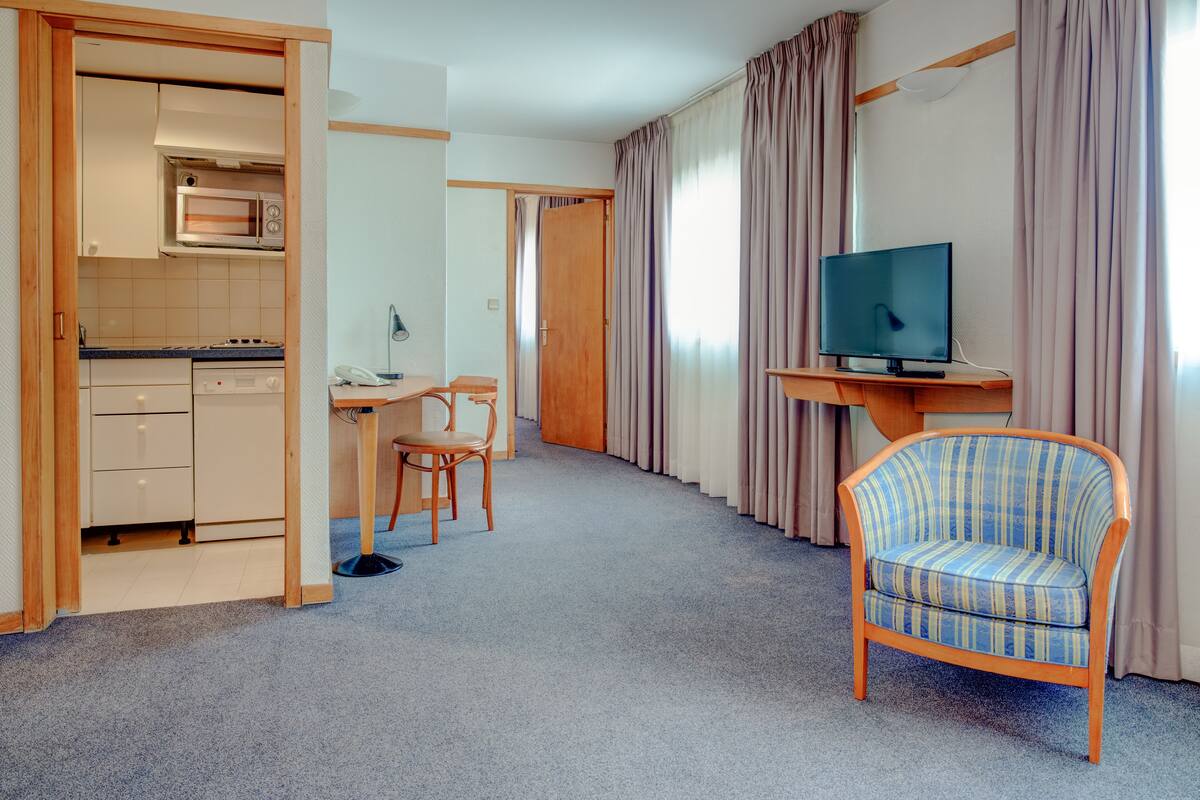 Ein kompaktes Hotelzimmer mit Küchenzeile, einem Essbereich, einem Fernseher und einem gestreiften Sessel.