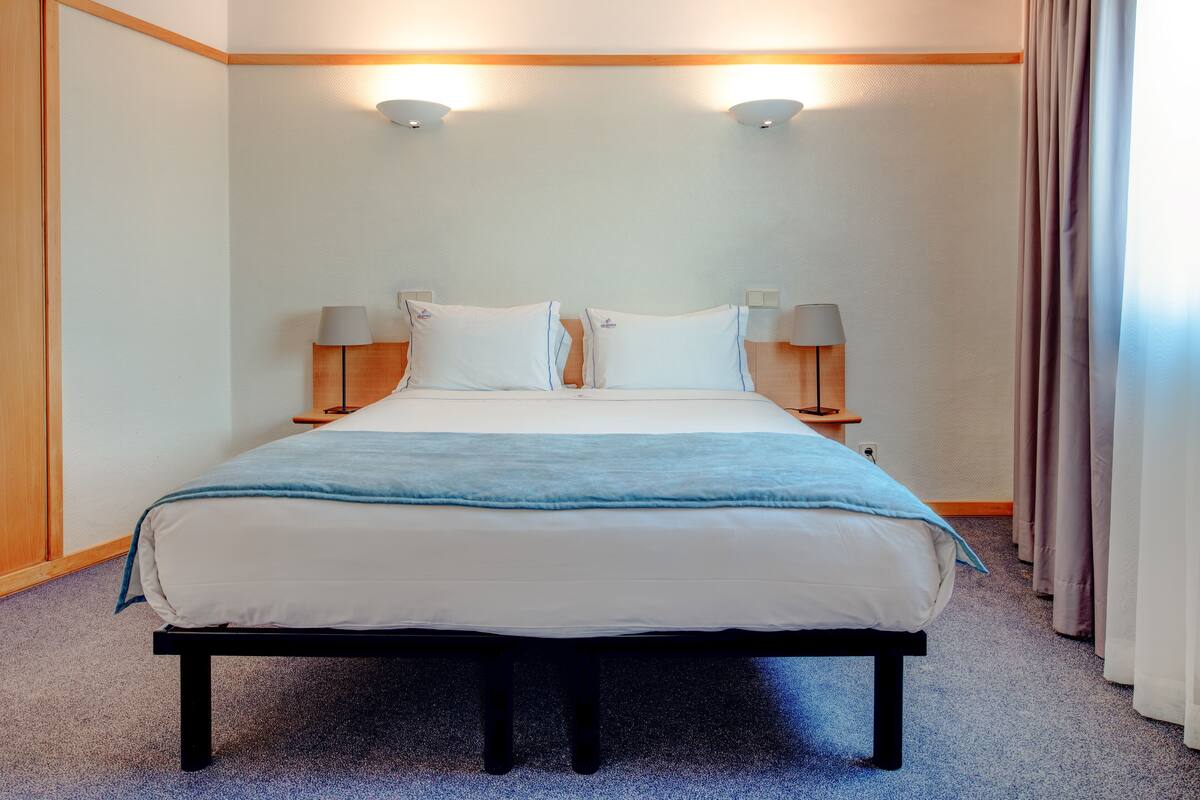Ein ordentlich bezogenes Bett mit blauer Decke, zwei Nachttischlampen und einem Kopfteil aus Holz.