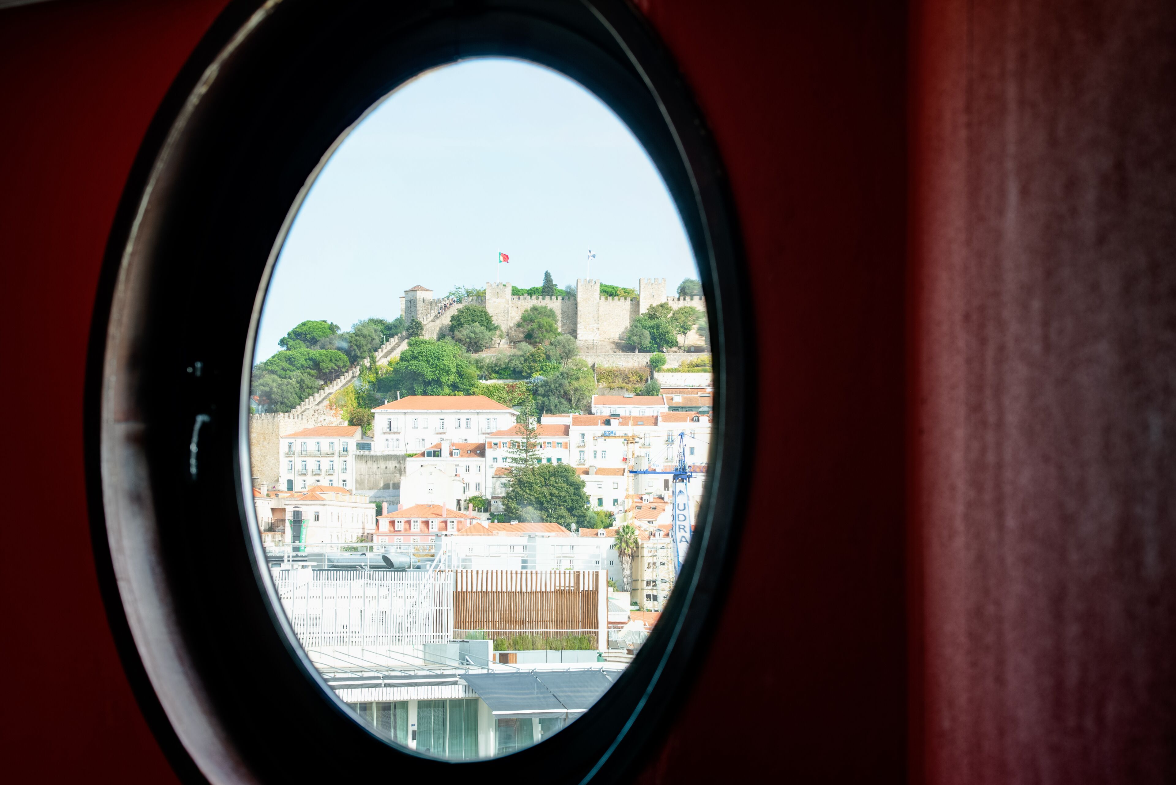 Vista a través de una ventana de un paisaje urbano con un castillo destacado en una colina, rodeado de edificios con techos de tejas rojas.
