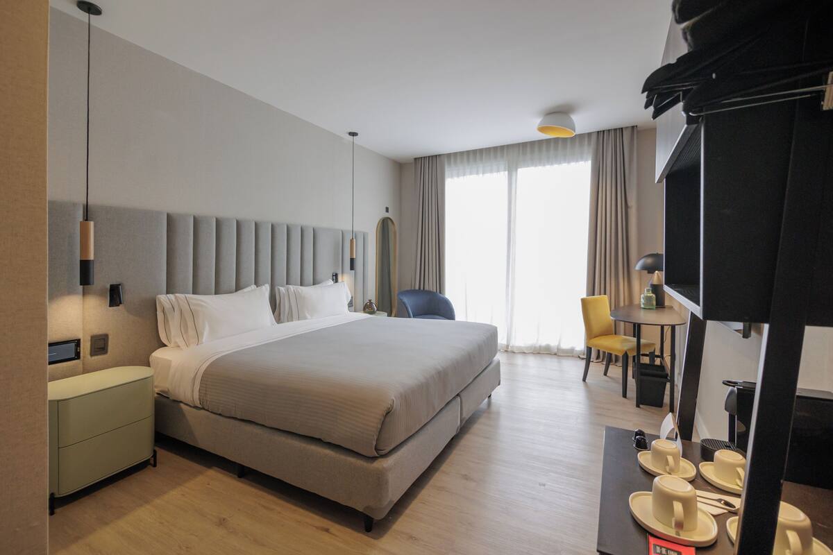 Una habitación de hotel con cama, mesitas de noche, un escritorio con silla, televisión y ventana con cortinas.