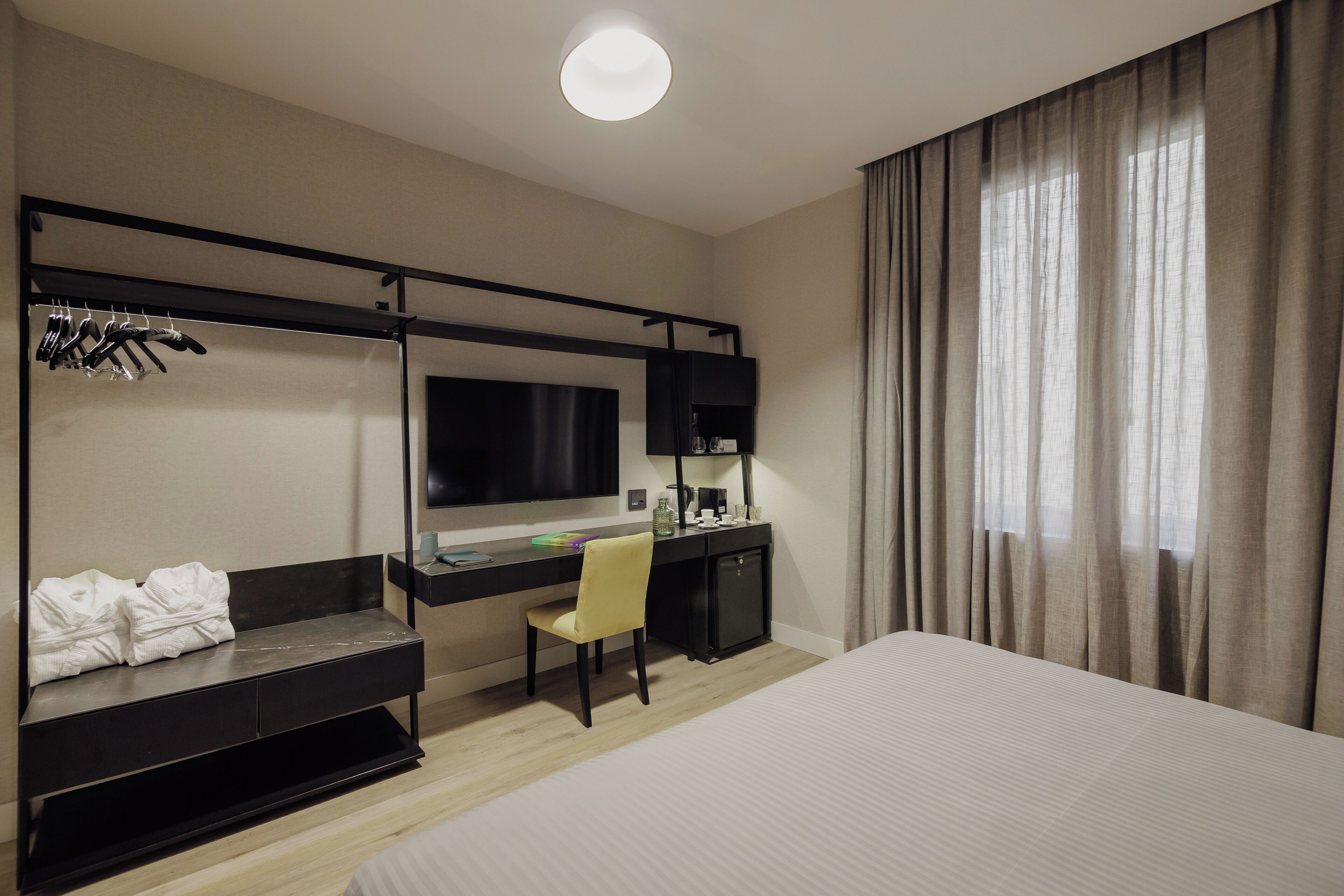A hotel room with a bed, a desk with a chair, a TV, and a wardrobe with hanging clothes.
