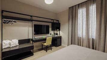 A hotel room with a bed, a desk with a chair, a TV, and a wardrobe with hanging clothes.