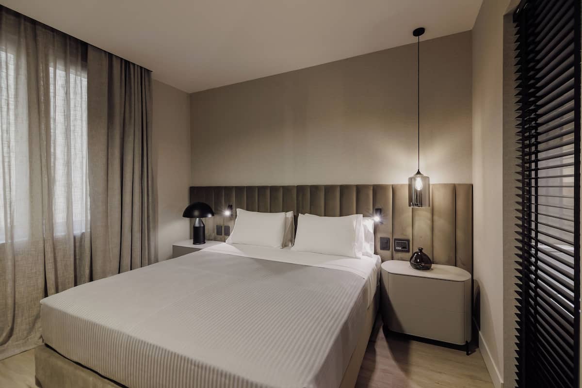 Habitación de hotel moderna con una cama grande, mesitas de noche, una lámpara y una ventana con persianas.