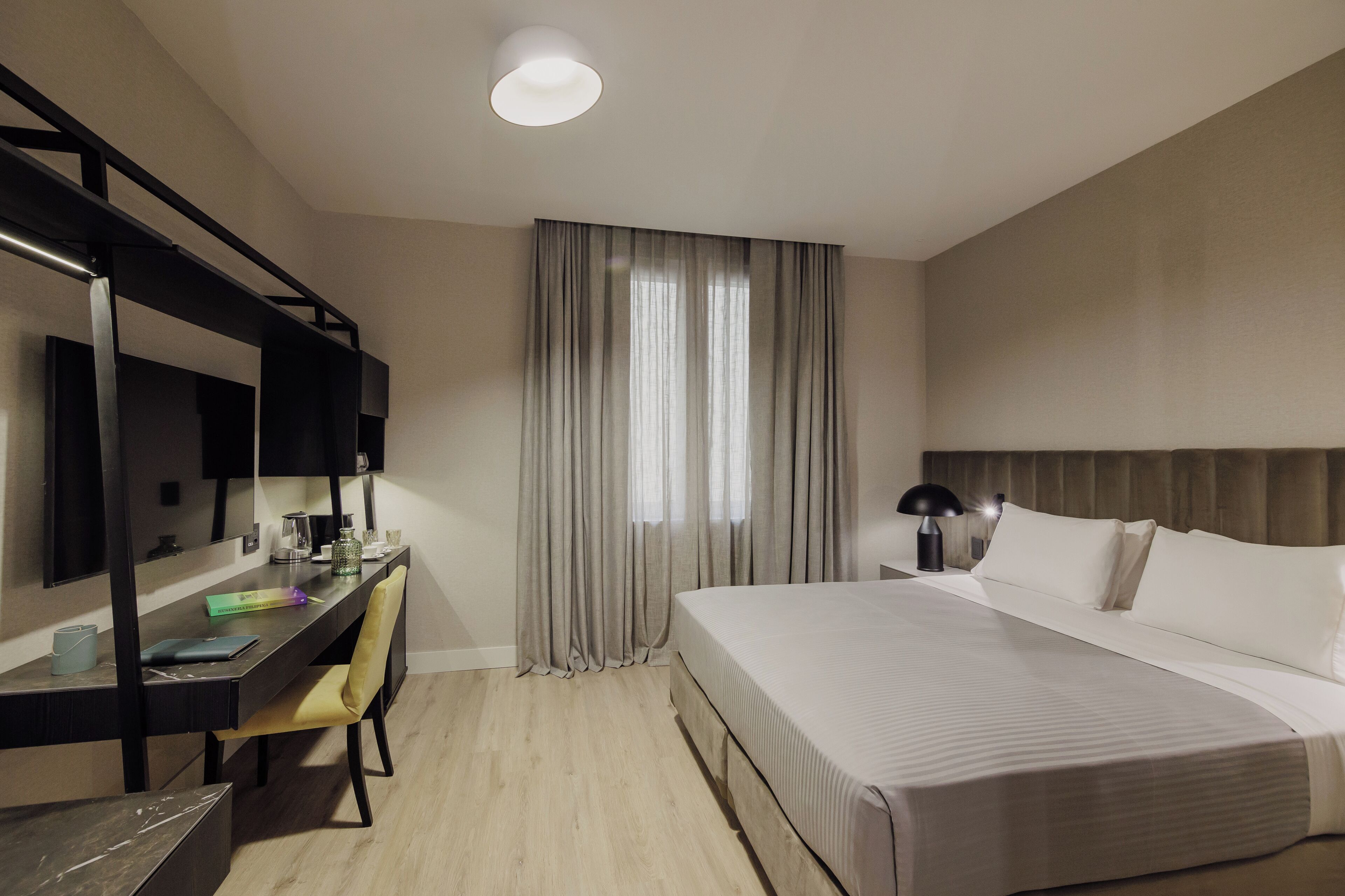 A hotel room with a bed, a desk with a chair, a mirror, and a window with curtains.