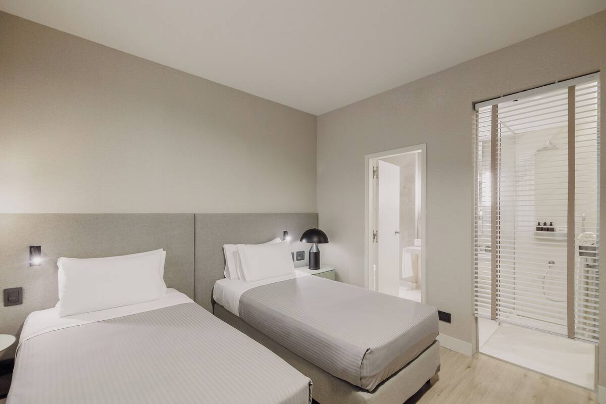 Una habitación de hotel moderna con dos camas, una mesita de noche, una lámpara y una puerta corrediza hacia el baño.