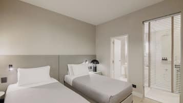 A modern hotel room with two beds, a nightstand, a lamp, and a sliding door to a bathroom.
