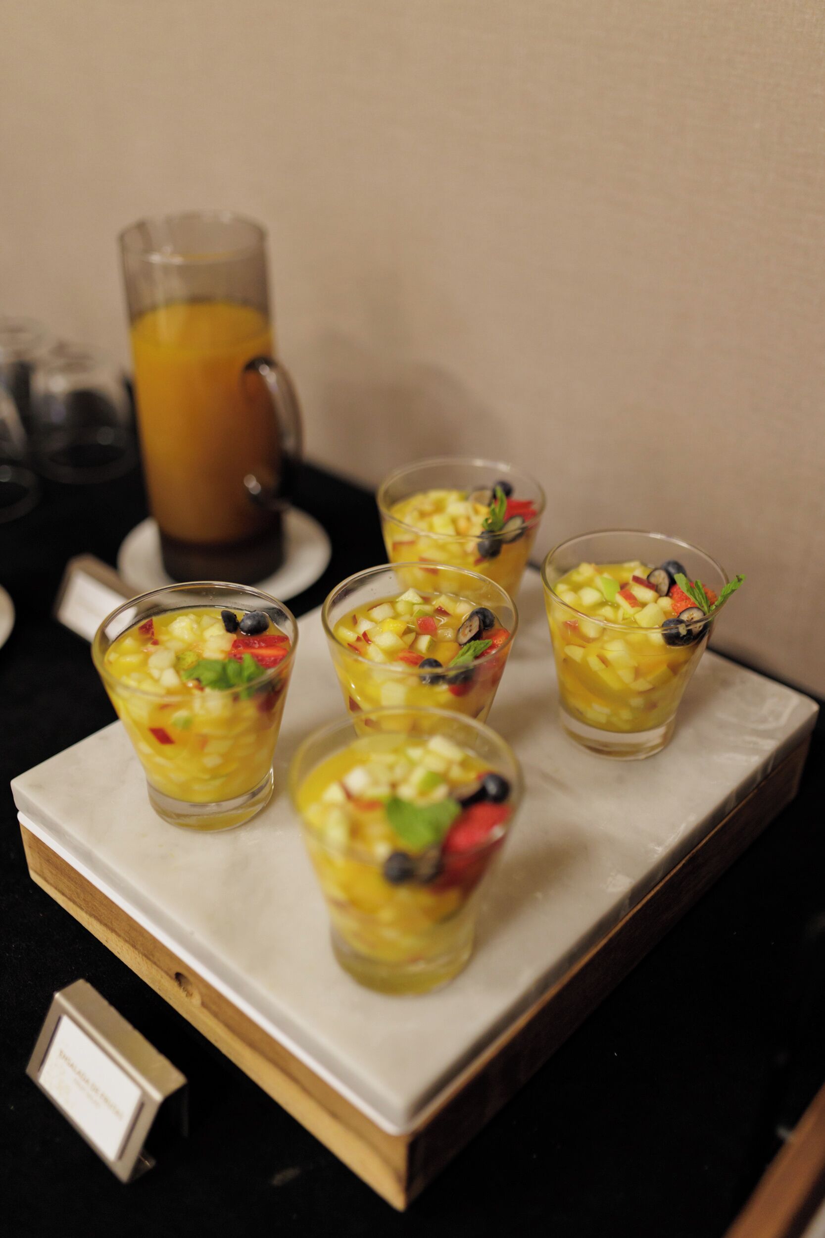 Un plateau contenant cinq verres de salade de fruits et une carafe de jus d’orange, posés sur une table.